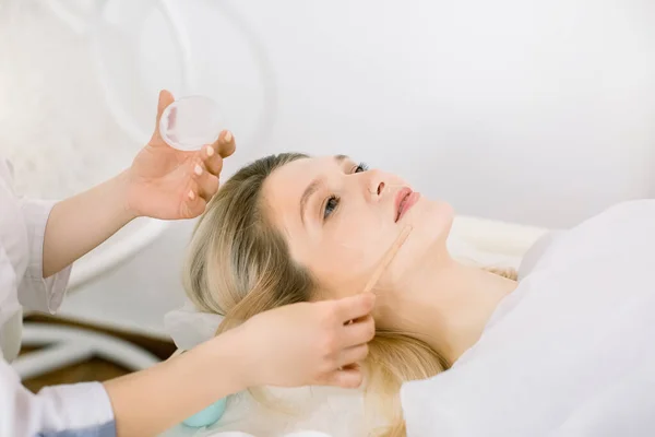 People, beauty, spa, cosmetology and skincare concept. Close up of beautiful young woman lying on the couch and female doctor cosmetologist applying facial mask by wooden spatula in spa center