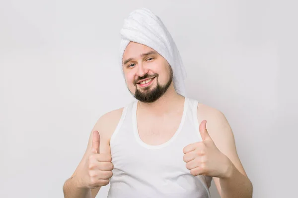 Ευτυχισμένος χαμογελαστός Καυκάσιος γενειοφόρος άντρας με λευκό μπλουζάκι και με μια πετσέτα στο κεφάλι να ποζάρει στην κάμερα σε απομονωμένο φόντο και να δείχνει τους αντίχειρές του ψηλά. Διαδικασίες Spa για τους άνδρες — Φωτογραφία Αρχείου