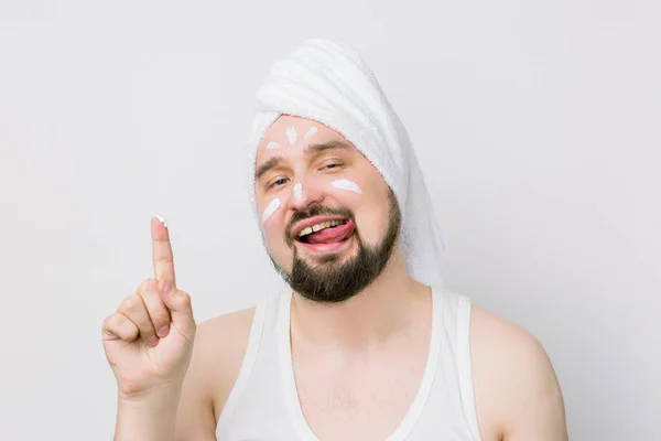 Funny bearded man with white towel on his head showing his finger with face cream on it, smears a cream on his face, smiling and showing his tongue. The concept of male self-care and male cosmetics — Stock Photo, Image