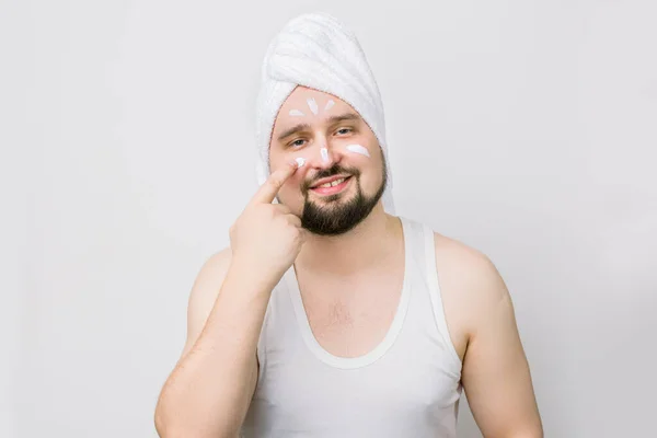 Funny smiling young caucasian man with mustache and beard, with white towel on head smiling at camera, smearing cream oh his face. Body or skin care for man health. Studio shot on white background. — Stock Photo, Image