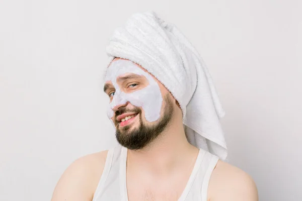 Portrait of a handsome joyful relaxed young bearded man with a facial mud clay mask, with towel on his head, posing with funny emotions on isolated white background. Face and body skin care treatment. — Stock Photo, Image