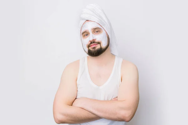 Handsome bearded man with white towel on his head and cosmetic mask on his face, stands with arms crossed on white background. Cosmetology procedures for men. Skin care for men. — Stock Photo, Image