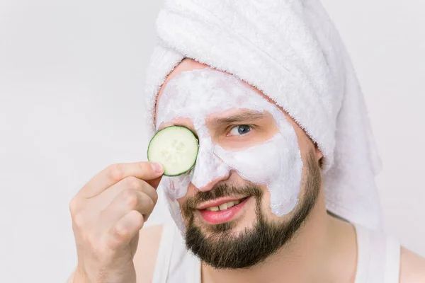 Zbliżenie portret nieogolonego brodatego mężczyzny w białym ręczniku och jego głowa, w kosmetycznej masce po prysznicu, pozowanie z plasterkiem ogórka zakrywającym jego oko — Zdjęcie stockowe