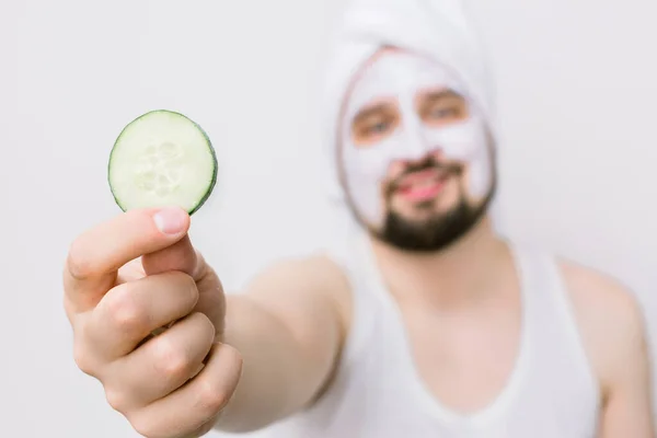 Blurred face of handsome bearded man in white towel having white mask on his face and posing with slice of cucumber, showing it to camera. Man with mask on face, white background, focus on cucumber — Stock Photo, Image