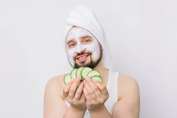 Close up portrait of happy funny bearded man in white towel with facial clay mask for skin care, having fun holding cucumber slices isolated over white background. Skin care for men, hygiene concept — Stock Photo, Image