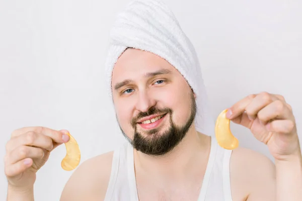 Young man wants to look fresher and caring about his skin. Handsome bearded guy in towel on head holds golden anti wrinkle eye patches, posing at camera with smile, isolated on white background — Stock Photo, Image