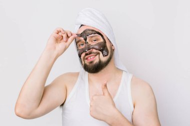 Erkekler için kaplıca ve cilt bakımı. Beyaz arka planda siyah akne kömür maskeli komik bir genç, maskesini çıkartıyor ve baş parmağını gösteriyor.