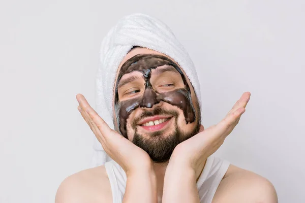 Smiling cheefrul Caucasian bearded man in towel on head, posing on white background, keeping face on his hands,enjoying skin care procedures. Acne removal with black charcoal mask, cleaning pores — Stock Photo, Image