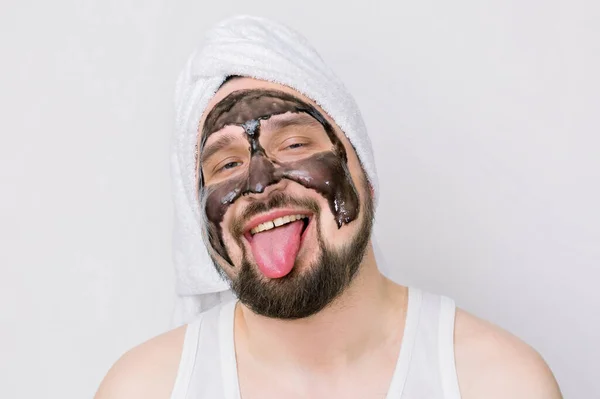 Um conceito divertido de cosmetologia. Homem barbudo engraçado com toalha branca oh sua cabeça, com máscara de creme preto em seu rosto, mostrando língua, estar de bom humor durante o tratamento de spa da manhã . — Fotografia de Stock