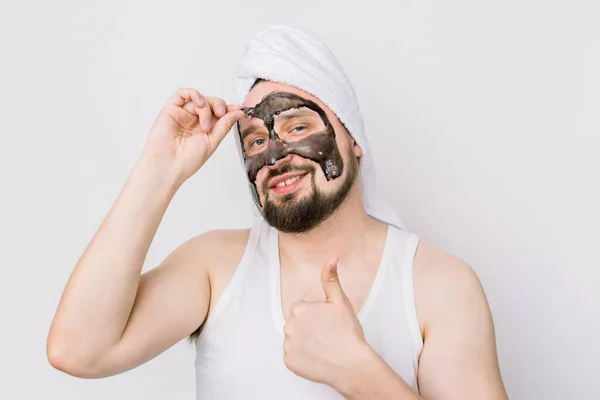 Spa and skin care for men. Young funny guy with black anti acne charcoal mask on his face on white background, removing mask from face and showing thumb up — Stock Photo, Image