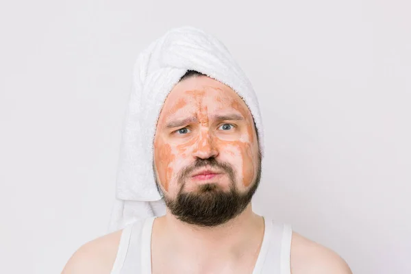 Funny bearded handsome man with white towel on head, with a brown mud mask on face, looking at camera on white background. Man with expressive face, copy space. Spa and skin care for men — Stock Photo, Image