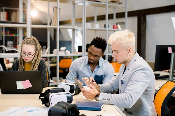 Bel homme concentré, travailleur d'affaires, assis dans la salle de bureau pendant la réunion et en utilisant la tablette ipad. Beau Africain homme et blond fille caucasienne avec dreadlocks travailler sur le fond — Photo