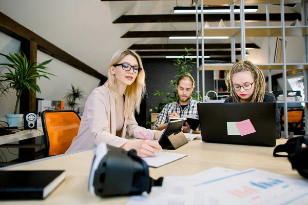 Grupo de jovens designers criativos, empresários, reuniram-se para discutir a ideia, desenvolver startup. Duas meninas bonitas e cara bonito trabalhando na mesa de escritório usando aparelhos eletrônicos — Fotografia de Stock