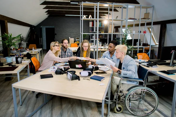 Życie biurowe, spotkanie biznesowe. Wielokulturowa grupa kreatywnych młodych współpracowników omawiających strategię biznesową, siedzących przy stole. Ładna blondynka daje papiery przystojnemu niepełnosprawnemu facetowi na wózku inwalidzkim — Zdjęcie stockowe