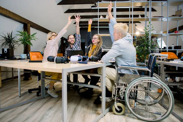 Pohled zezadu pohledného mladého zdravotně postiženého podnikatele na vozíčku se svými kreativními kolegy, oslavující úspěšný projekt se šťastnými úsměvy a zdviženými pažemi, sedícího u stolu v kancelářské místnosti — Stock fotografie