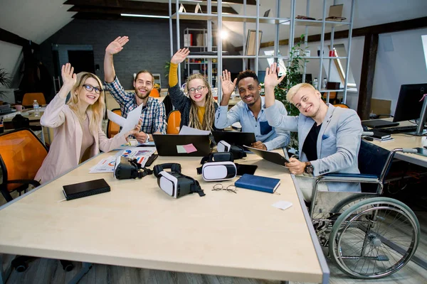Tým mnohonárodních obchodníků sedících během setkání u kancelářského stolu, dívajících se s úsměvem na kameru a mávajících rukama. Zařazení pro handicapovaného muže na invalidním vozíku — Stock fotografie
