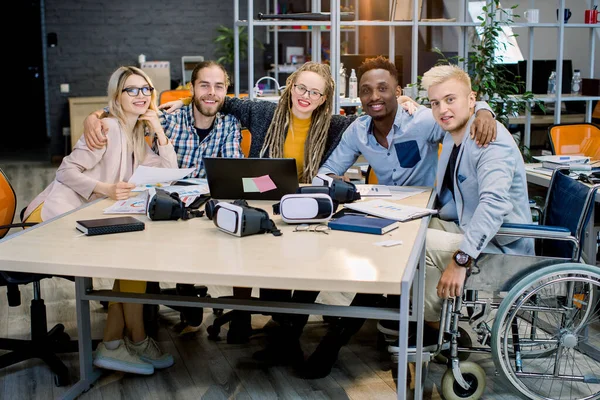 Inclusion dans le travail, concept d'équipe d'affaires. Beau collègue masculin en fauteuil roulant avec ses collègues d'affaires créatifs dans la salle de réunion au bureau créatif, souriant, regardant la caméra, s'embrassant — Photo