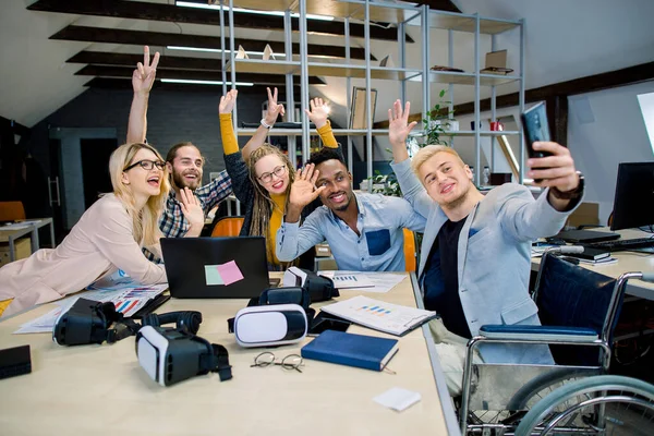 Snygg glad man i rullstol sitter vid skrivbordet med sina kollegor, och gör roliga selfie foto på smartphone. Grupp av multietniska hipster medarbetare poserar för foto och vinka — Stockfoto