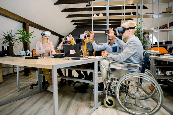 숙련 된 다종의 젊은 동료들을 만나 테이블에 앉아 Vr 고글을 사용하여 사무실에서 함께 일합니다. 휠체어에 앉아 창의적 인 사무실에서 친구들 과 함께 일하는 다루기 힘든 장애 인 — 스톡 사진