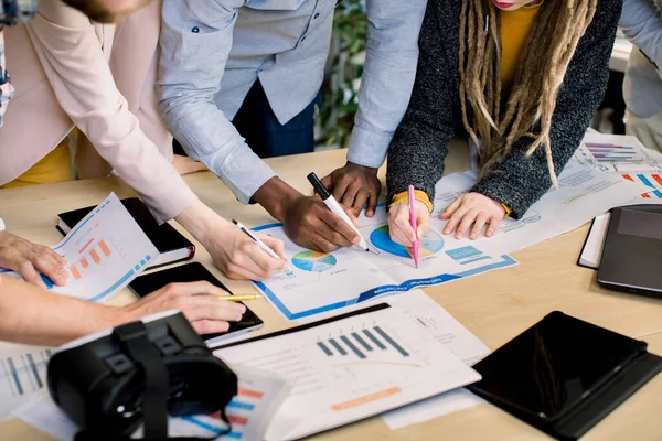 Image rapprochée d'une équipe d'affaires multiethnique diversifiée analysant des graphiques et des graphiques de revenu avec stylos dans les mains. Ordinateur portable moderne, tablette et lunettes vr sur la table. Concept de stratégie — Photo