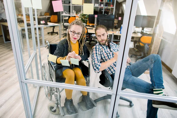 Jeune femme d'affaires hipster handicapée souriante avec dreadlocks blonds, assise en fauteuil roulant, lisant des notes collantes sur un mur de verre et écrivant ses idées tout en faisant du remue-méninges avec un collègue masculin au bureau — Photo