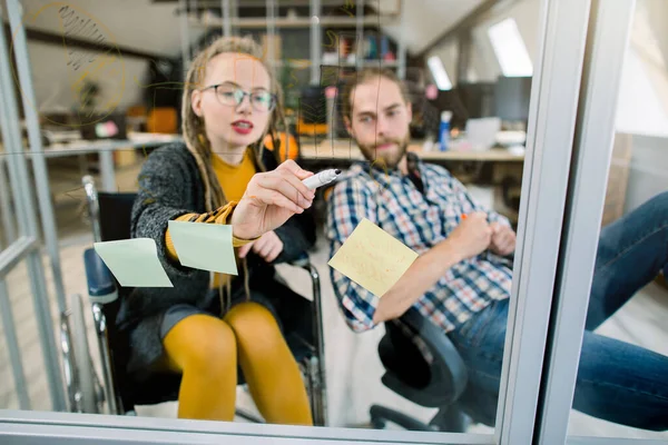 Group of young successful creative team, skilled handicapped blond girl with dreadlocks, sitting in wheelchair, and handsome bearded man, brainstorm on project together in modern office
