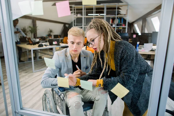 Deux hommes d'affaires heureux créatifs concentrés, homme handicapé en fauteuil roulant et jolie fille blonde avec dreadlocks, remue-méninges au bureau, plaçant des notes collantes avec des idées sur le mur de verre — Photo