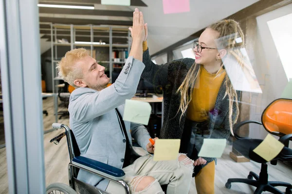 Business concept, successful disabled people at work. Happy successful young man in wheelchair giving a high five to his pretty female coworker with dreadlocks, laughing and cheering their success
