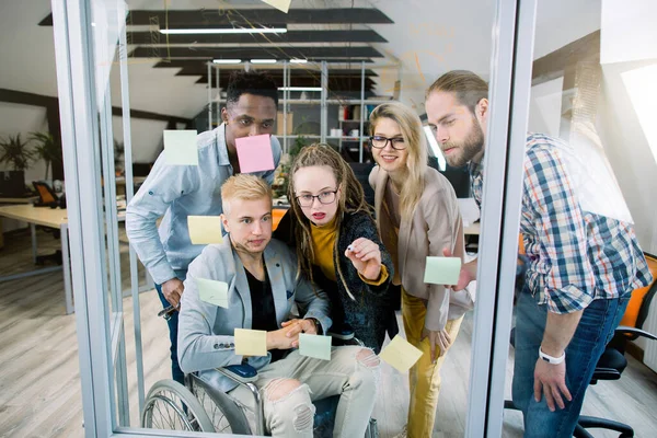 Integration för funktionshindrade. Grupp unga multiraciala affärsmän med en handikappad man i rullstol, diskuterar idéer framför glasväggen med hjälp av post det anteckningar och klistermärken — Stockfoto