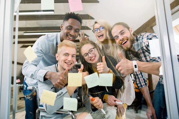 Divers groupes d'hommes d'affaires multiethniques souriants faisant du remue-méninges avec des notes collantes sur un mur de verre tout en travaillant dans un bureau moderne, regardant la caméra à travers le verre et montrant les pouces vers le haut — Photo