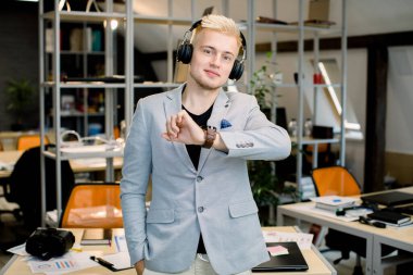 Yakışıklı, kendine güvenen beyaz erkek yönetici, kulaklık takıyor, kol saatine bakıyor, kameraya poz veriyor, modern yaratıcı parlak ofiste duruyor. Ofiste kulaklıklı bir iş adamı.