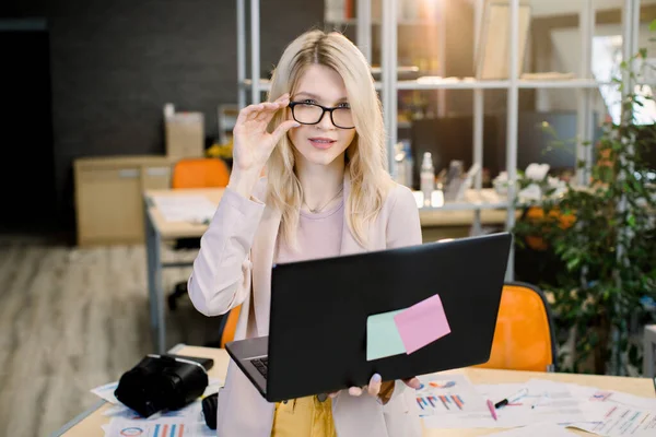 Gyönyörű fiatal szőke üzletasszony szemüvegben, okos alkalmi ruhákat visel, a kamerába néz és mosolyog, miközben a modern kreatív irodában dolgozik, laptopot használva — Stock Fotó