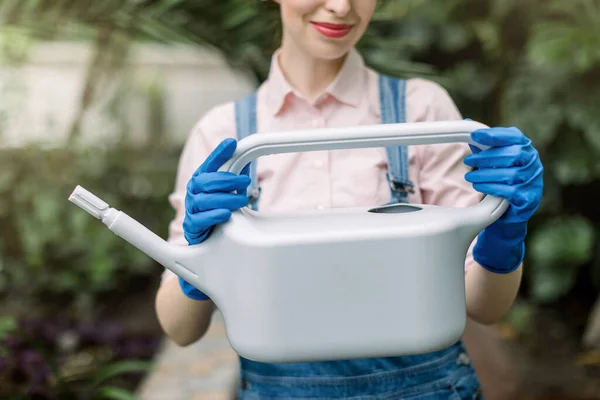 Immagine ritagliata di bella fiorista donna in tuta jeans e guanti di gomma blu, con in mano lattina d'acqua grigia per innaffiare le piante nella sua serra. — Foto Stock