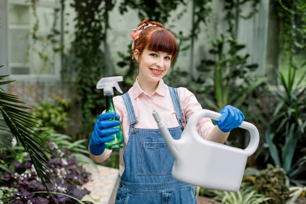 Giovane bella giardiniera femminile, indossando tuta jeans, posa alla macchina fotografica che tiene annaffiatoio e spruzzatore di plastica verde per le piante, in una serra. Concentrati sul volto della donna — Foto Stock