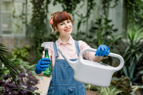 Giovane bella giardiniera femminile, indossando tuta jeans, posa alla macchina fotografica che tiene annaffiatoio e spruzzatore di plastica verde per le piante, in una serra. Concentrati sul barattolo e sull'irrigatore — Foto Stock