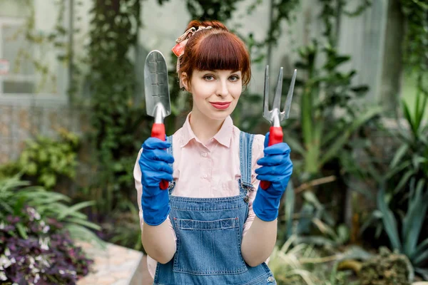 Ritratto di una giovane donna giardiniera in tuta jeans e guanti di gomma blu, in posa con attrezzi da giardinaggio all'interno in serra sullo sfondo di piante tropicali e succulente — Foto Stock