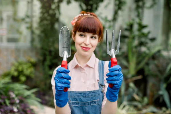 Ritratto da giardiniera. Ragazza dai capelli rossi sorridente, operaia di serra, in tuta jeans e camicia rosa, tenendo in mano attrezzi da giardinaggio e guardandolo. Concentrati su vanga e rastrello — Foto Stock