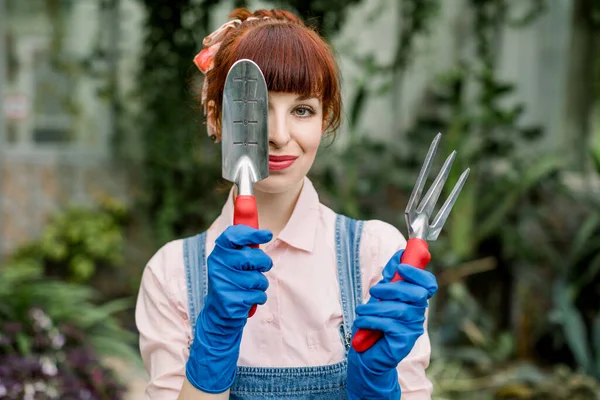 温室和种植概念。漂亮的红头发少女，在温室里干活，拿着园艺工具站着，摆姿势拍照，用铁锹遮住眼睛. — 图库照片