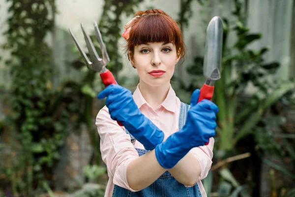 Grave giovane donna concentrata in guanti protettivi blu tenendo attrezzi da giardinaggio a mani incrociate e guardando la fotocamera. Trapianto di giardinaggio di concetto vegetale — Foto Stock