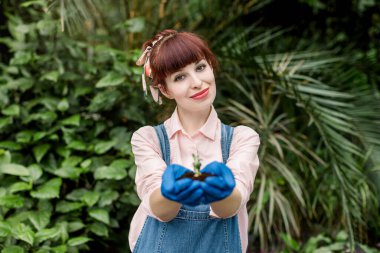 Kot pantolonlu, gülümseyen, sevimli kadın bahçıvanın elinde toprakla sulu bir bitki tutması. Serada fidan tutan kadın, kameraya bakıyor.