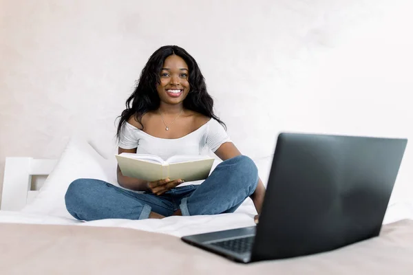 Giovane ragazza afroamericana in t-shirt bianca e jeans, seduta sul letto con pc portatile e blocco note in camera da letto, studiando o lavorando a casa, trascorrendo del tempo a casa. Concetto di lavoro online — Foto Stock