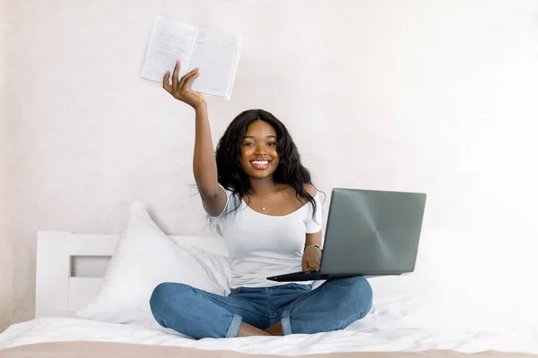 Giovane donna afroamericana in t-shirt bianche seduta sul letto con computer portatile in camera da letto, tenendo il taccuino che mostra alla fotocamera, sorridendo e divertendosi a lavorare e studiare a casa — Foto Stock