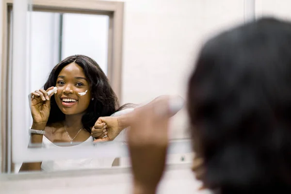 Reflection of pretty smiling African girl in mirror applying cosmetic cream on her face. Female putting moisturizer on her facial skin in light home bathroom. Spa and beauty at home