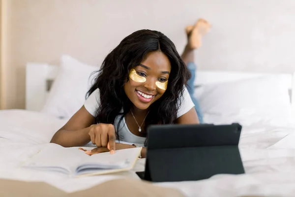 Sorridente donna africana si trova sul letto, scorrendo attraverso il suo computer tablet, lavorando online, facendo i compiti, preparando test o rilassarsi. Donna d'affari di successo con patch per gli occhi che lavorano su tablet — Foto Stock
