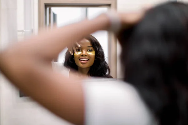 Tratamiento de belleza en casa por la mañana. Mujer africana cerca del espejo haciendo tratamientos de belleza en la cara, poniendo parches de ojos dorados en el área debajo de los ojos . —  Fotos de Stock