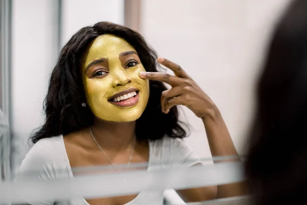 Pretty African woman looking in the mirror in bathroom, applying gold facial mask, touching her face with hands and looking at camera. Beauty, skin care and treatment peeling