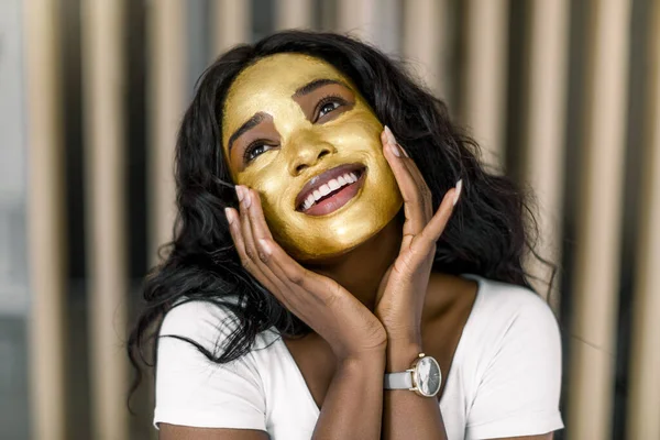 Spa, pielęgnacja skóry, kobiece leczenie twarzy. Piękno zbliżenie portret radosnej afrykańskiej kobiety ze złotym zdjęciem maski na twarzy dotykając jej twarzy i uśmiechając się, patrząc w górę. Przytulne wnętrze domu — Zdjęcie stockowe
