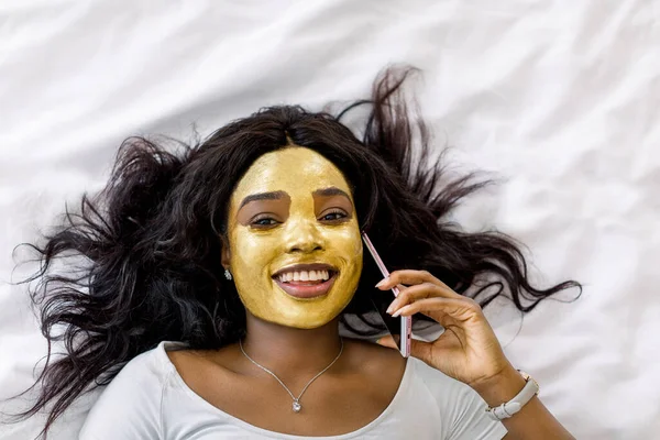 Vista superior de la encantadora mujer africana sonriente con máscara facial de arcilla dorada acostada en la cama y usando el teléfono celular, hablando con su amiga, mientras disfruta del tratamiento del cuidado de la piel —  Fotos de Stock