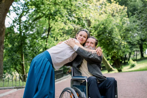 Disabled people, palliative care. Lifestyle summer portrait of senior bearded man in wheelchair hugging his young pretty hipster granddaughter, walking in park outdoors