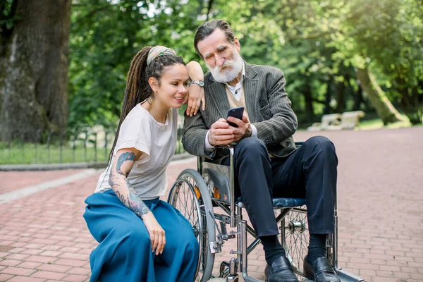 Porträt einer glücklichen Hipster-Enkelin mit Dreadlocks und einem barbusigen Großvater im Rollstuhl, der an einem sonnigen Tag in einem grünen Park Spaß hat, Fotos macht oder im Internet telefoniert. — Stockfoto